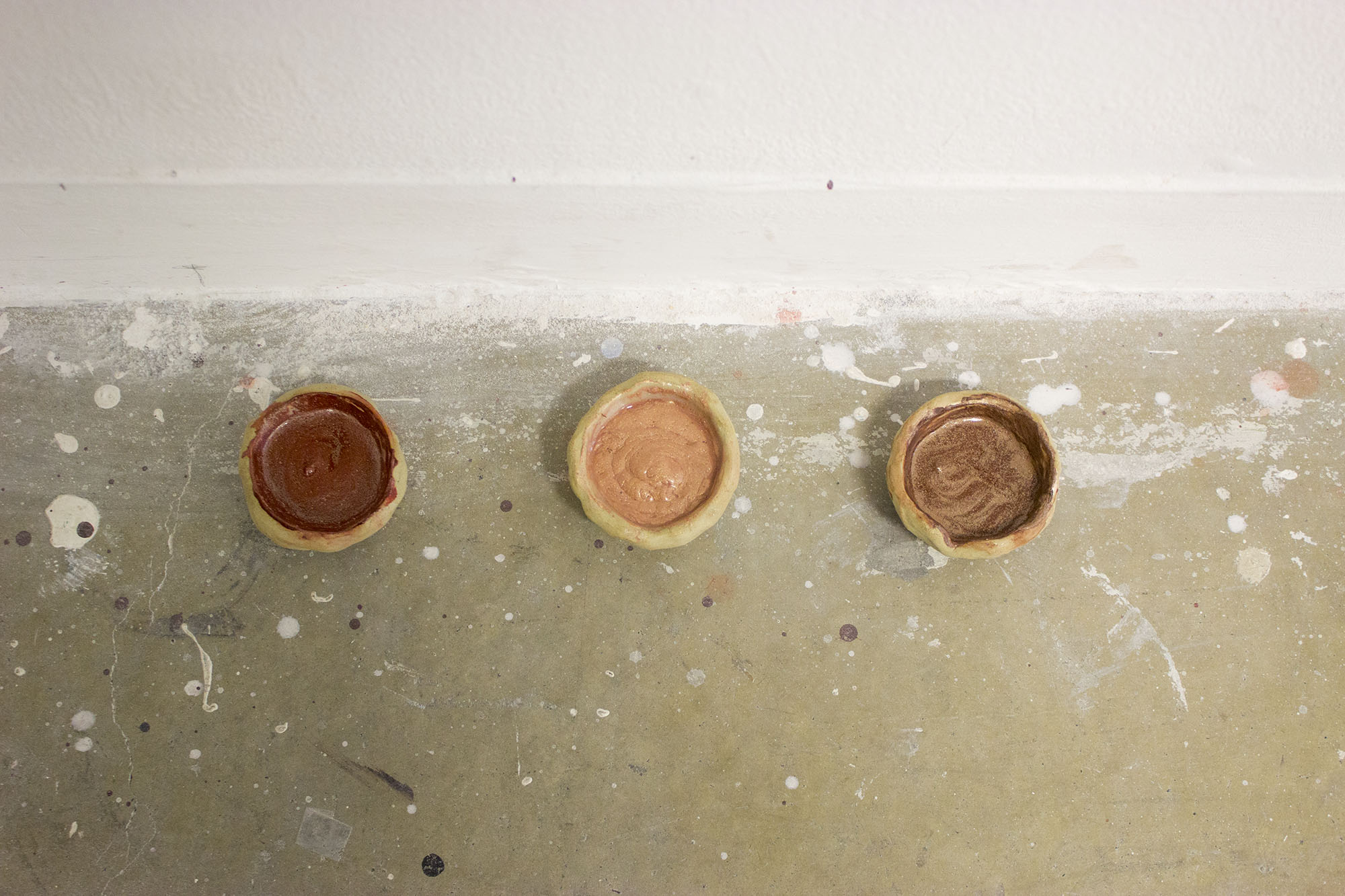 trois pots de peinture en terre posés au sol, on dirait des petits bols. Les peintures sont dans les tons ocre et sont metallisées.